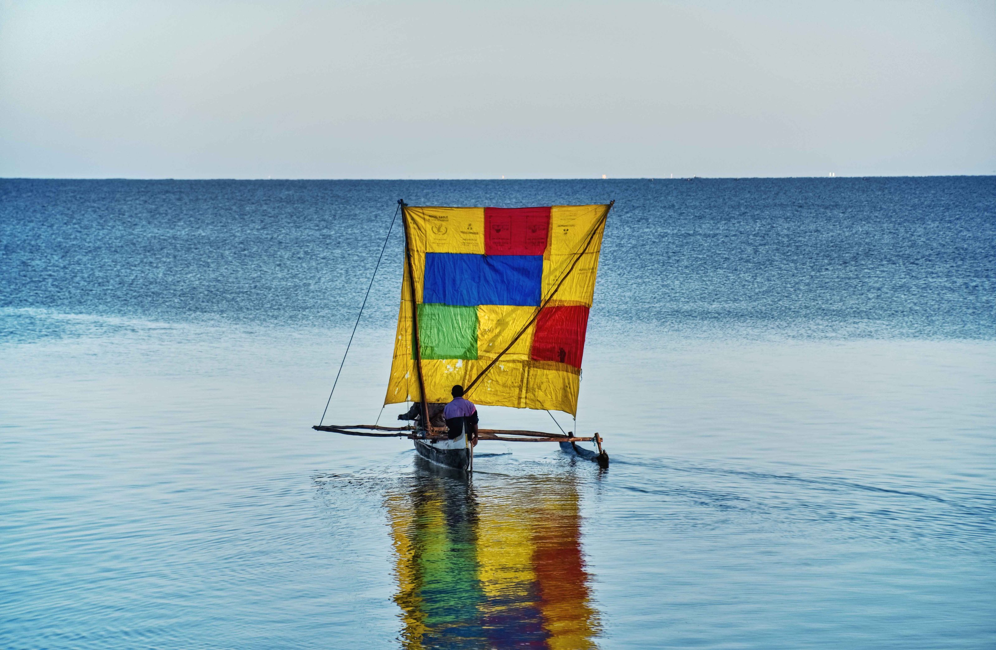 Une pirogue à balancier à la voile rapiécée jaune, rouge, bleue et verte, sur une eau calme et bleue, à Madagascar.